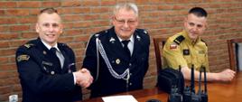 Siedzący za stołem Zastępca Komendanta Centralnej Szkoły PSP w uścisku dłoni z siedzącym obok druhem OSP obok siedzący funkcjonariusza wydziału informatyki i łączności CS PSP. Przed nimi na blacie przekazywany sprzęt łączności radiowej