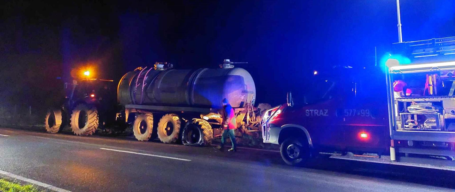 Kolizja na drodze wojewódzkiej.Na prawym pasie ruchu stoi ciągnik rolniczy z podczepionym beczkowozem. Za nim znajduje się wóz straży pożarnej, który ma włączone niebieskie światła błyskowe i otwartą skrytkę na sprzęt. Na dachu ciągnika włączone jest pomarańczowe światło ostrzegawcze. Pomiędzy beczkowozem a wozem strażackim znajduje sie mężczyzna.