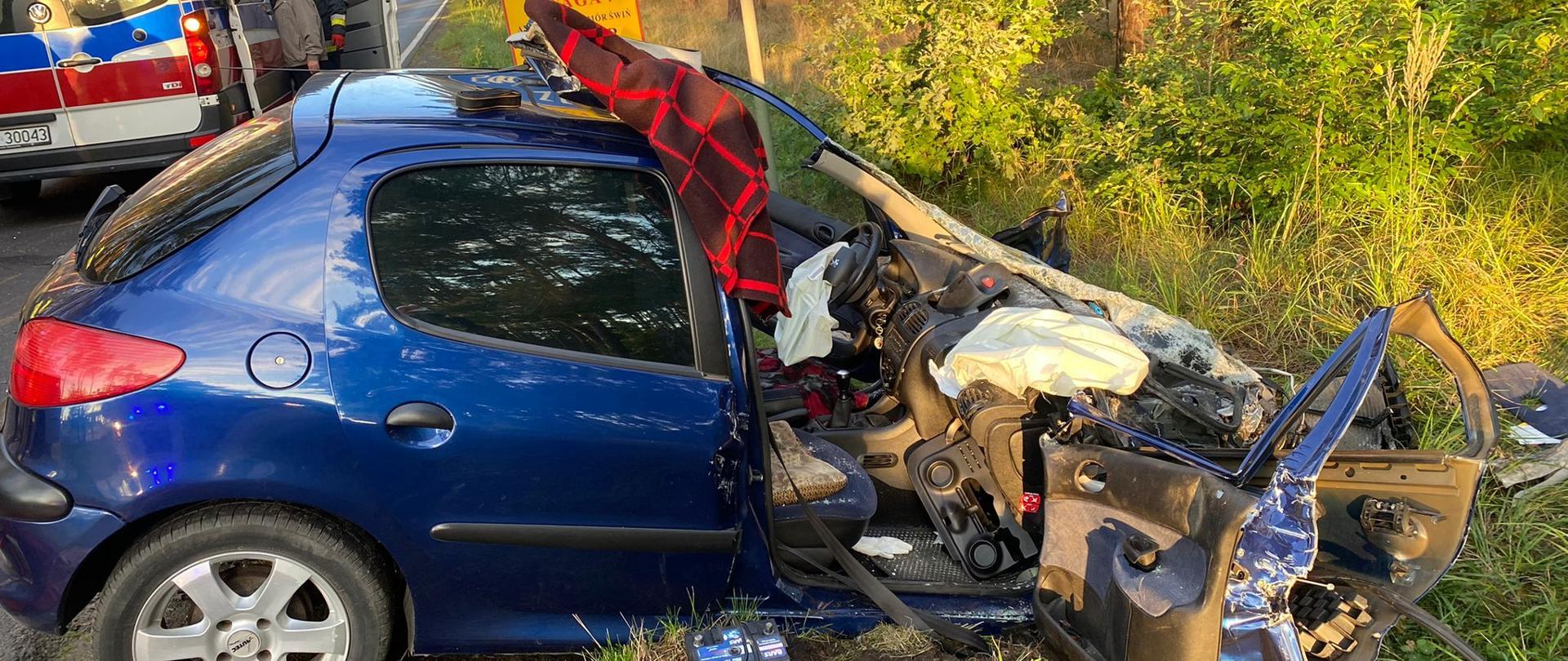 Dzień. Rozbity samochód Peugeot, w tle karetka pogotowia ratunkowego.