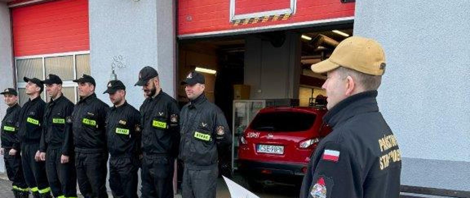 Strażacy w ubraniach koszarowych stoją w szeregu przed budynkiem Komendy Powiatowej Państwowej Straży Pożarnej w Sępólnie Krajeńskim, w tle garaże Jednostki Ratowniczo-Gaśniczej, na środku Komendant Powiatowy starszy brygadier Maciej Suchomski w ubraniu służbowym.