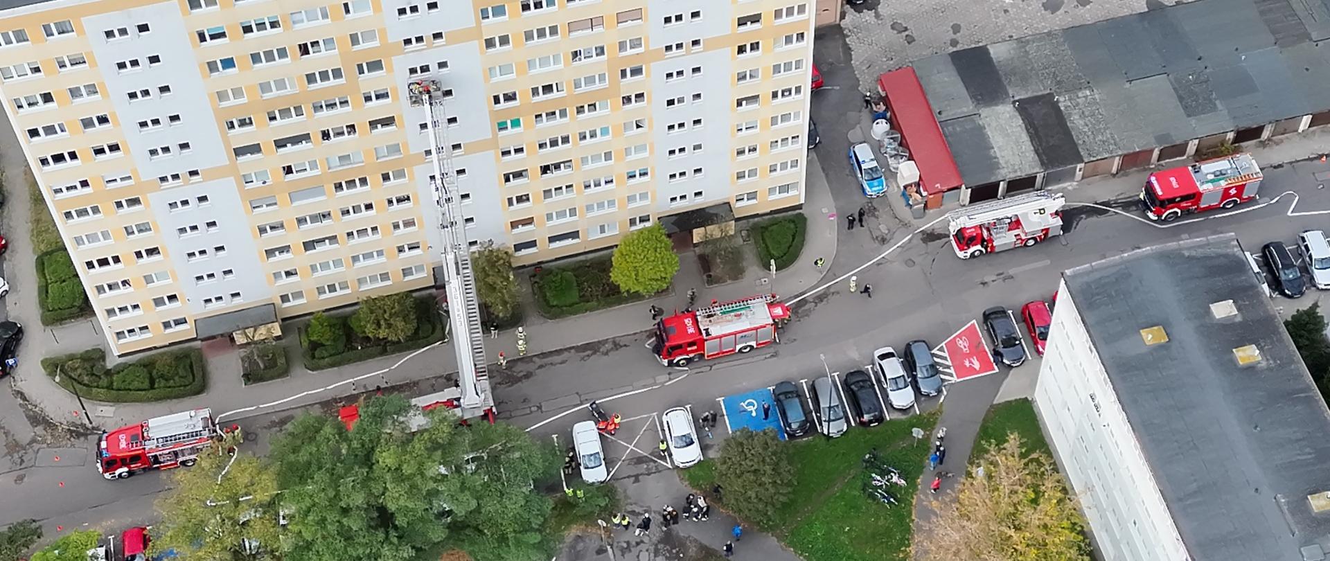 Widok z góry na teren ćwiczeń przy wieżowcu. Przy elewacji rozstawiona podnośnik hydrauliczny, wokół niego znajdują się wozy strażackie, karetki i uczestnicy ćwiczeń.