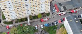 Widok z góry na teren ćwiczeń przy wieżowcu. Przy elewacji rozstawiona podnośnik hydrauliczny, wokół niego znajdują się wozy strażackie, karetki i uczestnicy ćwiczeń.