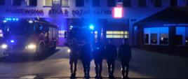 Strażacy oddają hołd śp. sekc. Bartosza Błyskala. Stoją salutując na tle samochodów pożarniczych z włączonymi sygnałami świetlnymi i dźwiękowymi.