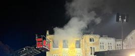 Zdjęcie w porze nocnej. Widać pomieszczenia objęte pożarem. Ściana kamienicy oświetlona sztucznym światłem. Występuje zadymienie.