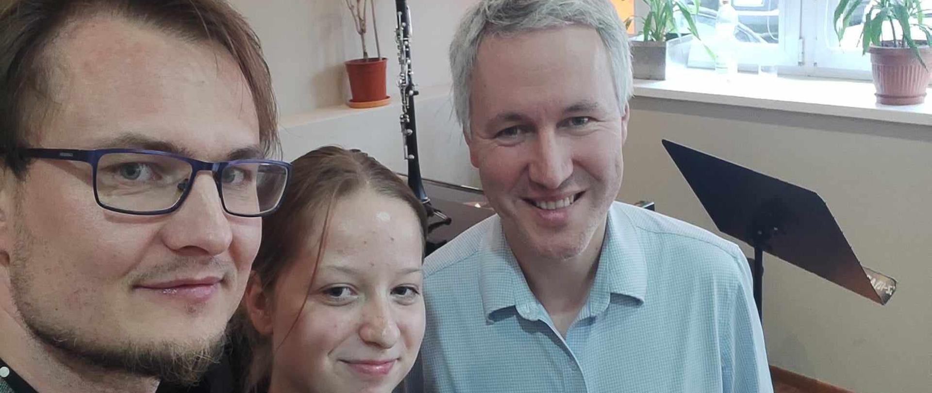 Zdjęcie przedstawia portret nauczyciela po lewej uczennicy w środku i nauczyciela po prawej stronie. Uczennica trzyma w ręku klarnet. W tle widać biurko i klarnet stojący na stojaku oraz okno z kwiatkami na parapecie.