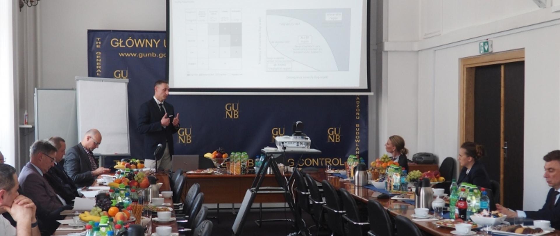 Osoby siedzące przy długim stole konferencyjnym podczas prezentacji w GUNB; na ekranie widoczne wykresy i grafy, w tle adres www.gunb.gov.pl.