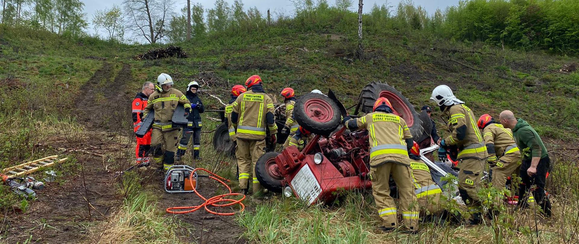 Na dole wzniesienia porośniętego zielonymi krzakami, czerwony traktor na dachu, widoczny przód, do góry kołami. W okół pracujący strażacy.