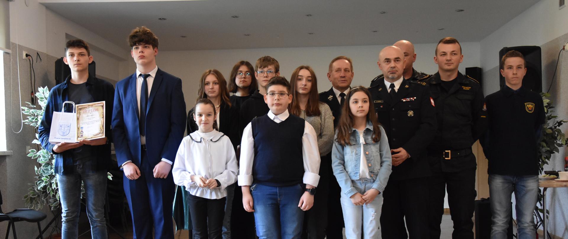 Na zdjęciu widzimy uczniów oraz strażaków, którzy pozują do zdęcia grupowego. Laureaci trzymają w ręku dyplomy za zajęte miejsca