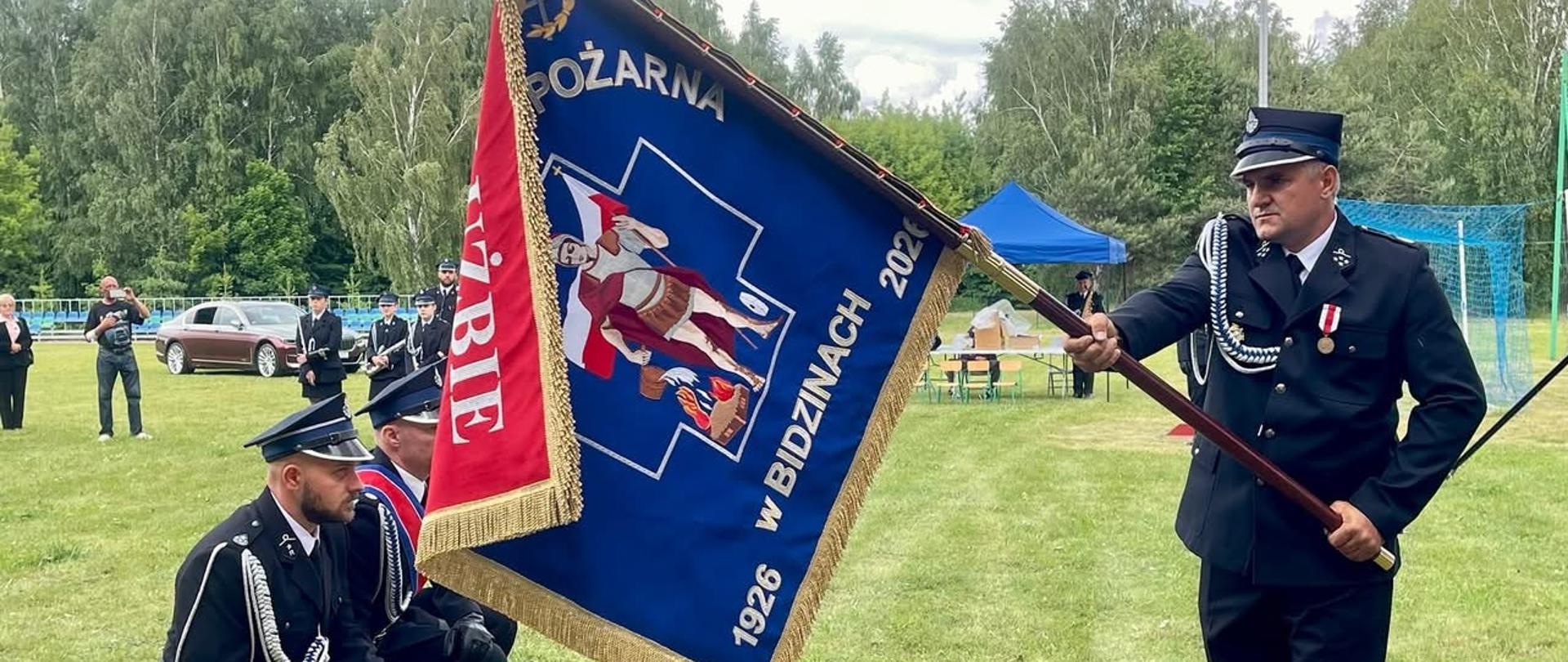 3 strażaków klęczy przed sztandarem trzymanym przez Prezesa OSP. Prezes OSP trzyma sztandar do prezentacji. Strażacy ubrani w mundury wyjściowe OSP