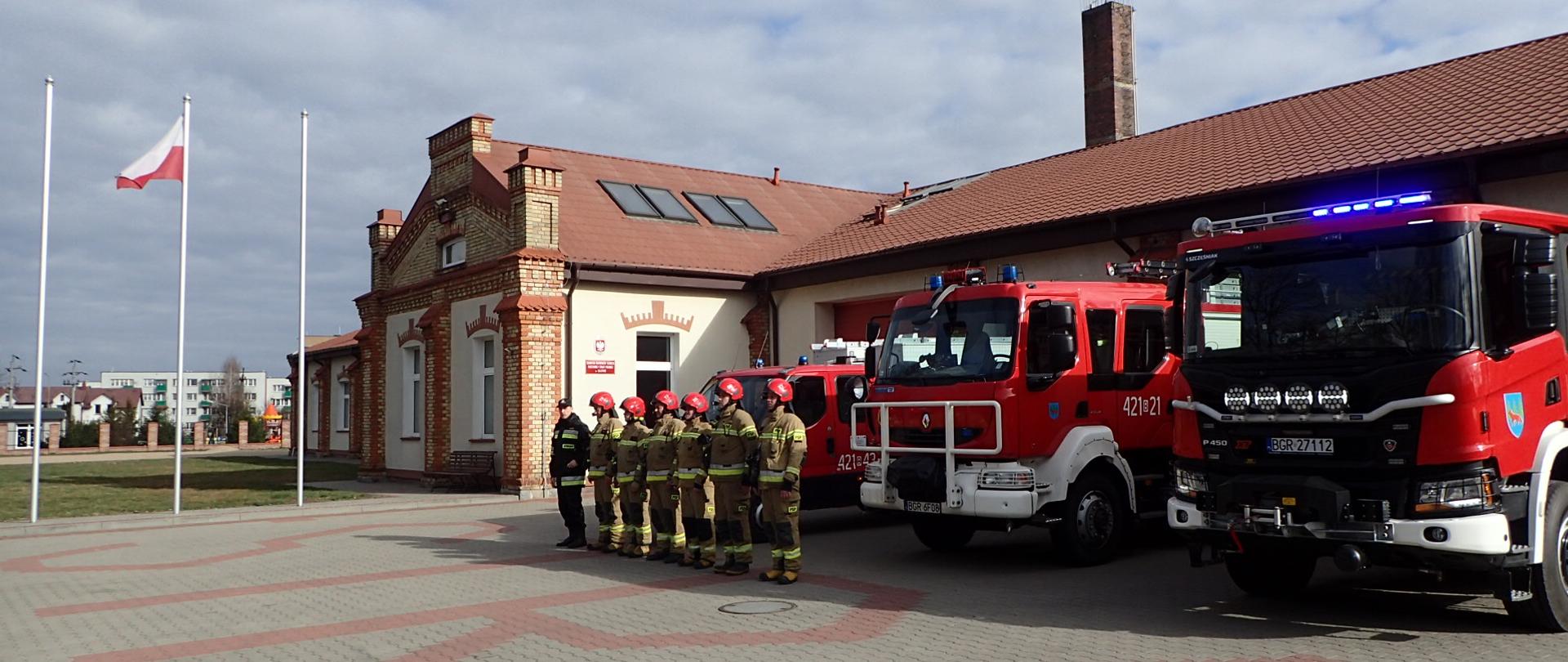 Strażacy PSP stoją na zbiórce przed strażnicą. Za nimi pojazdy strażackie. Z lewej w tle flaga polski powiewa na maszcie.