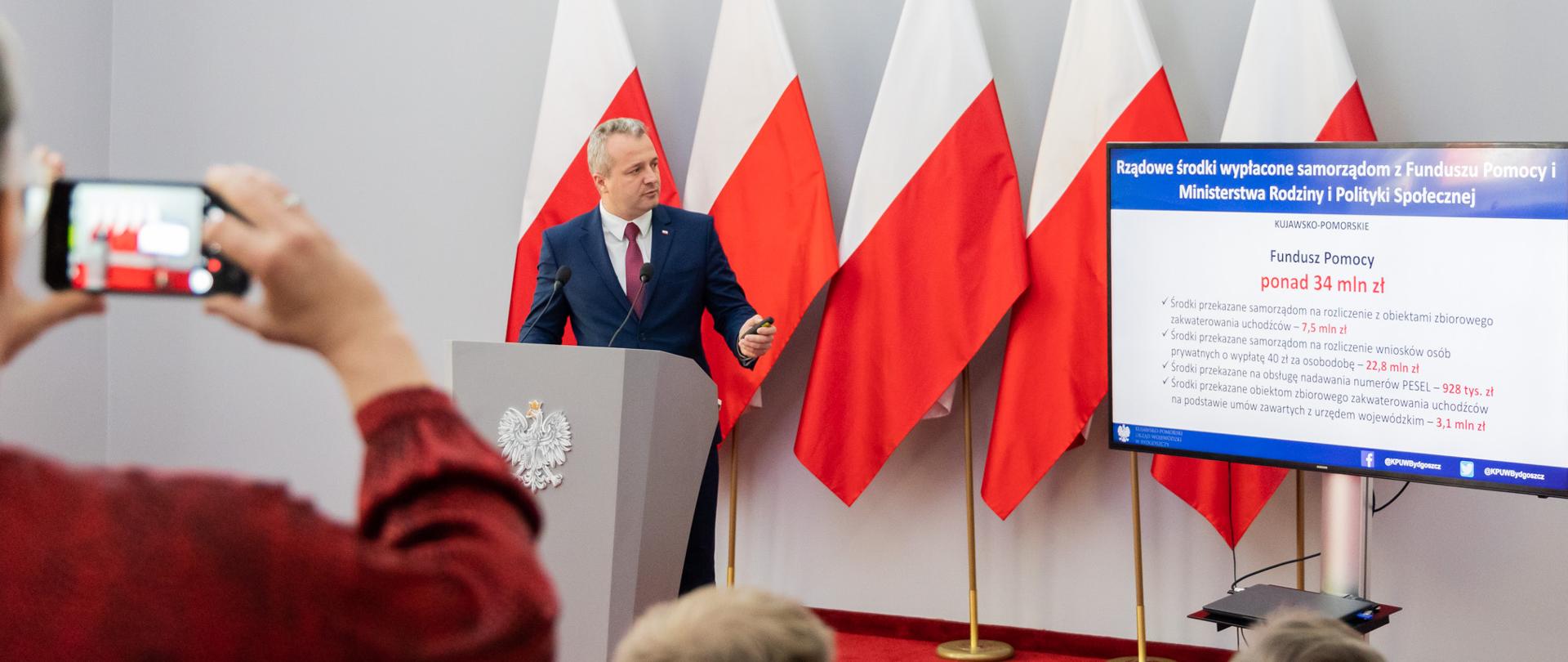 Jeden mężczyzna stojący przed mównicą i wypowiadający się. Przed nim siedzi kilka osób