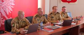 Czterech strażaków ubranych w służbowe mundury koloru piaskowego siedzi przy stole konferencyjnym, na którym znajdują się laptopy, dokumenty i kubki z kawą. W tle czerwony baner z orłem i napisem „Państwowa Straż Pożarna Komenda Powiatowa PSP w Świebodzinie”.