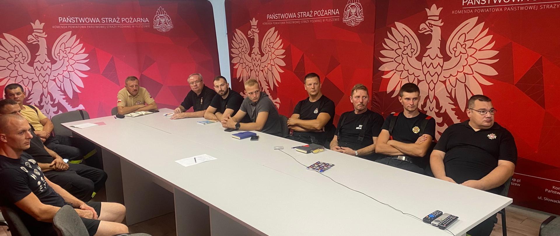 Grupa mężczyzn siedzi przy białym stole, w tle znajdują się banery straży pożarnej, na których widoczny jest orzeł państwowy oraz logo PSP i napis Państwowa Straż Pożarna na czerwonym tle.