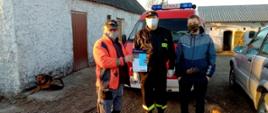 Zdjęcie przedstawia strażaka OSP, który wręczył dwóm mężczyzną ulotkę dotyczącą szczepień przeciwko COVID-19. Zdjęcie wykonane jest na tle samochodu strażackiego. Na zdjęciu widać dwa psy. W lewej stronie zdjęcia oraz w tle widać budynki gospodarcze. Po prawej stronie zdjęcia znajduje się samochód osobowy. 