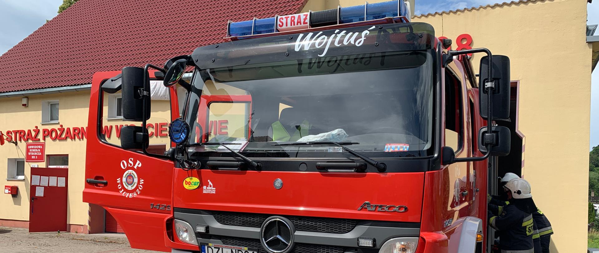 czerwony pojazd pożarniczy na tle remizy, po prawej stronie pojazdu umundurowania strażacy