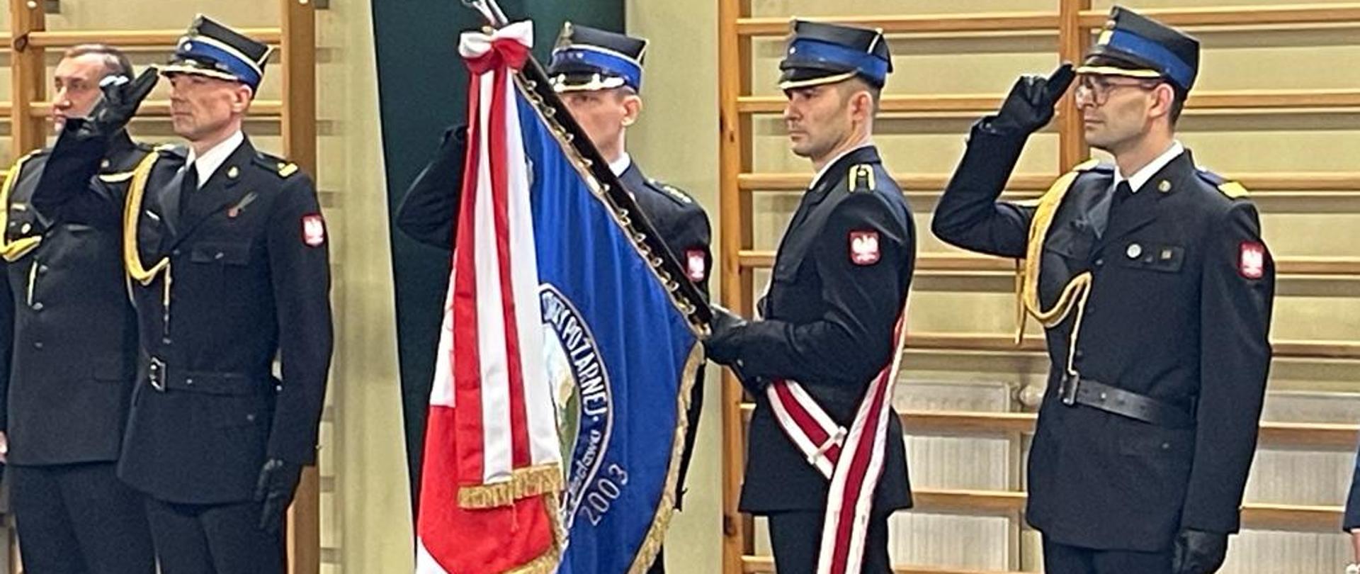 Strażacy stojąc podczas apelu, oddając honory w umundurowaniu wyjściowym.