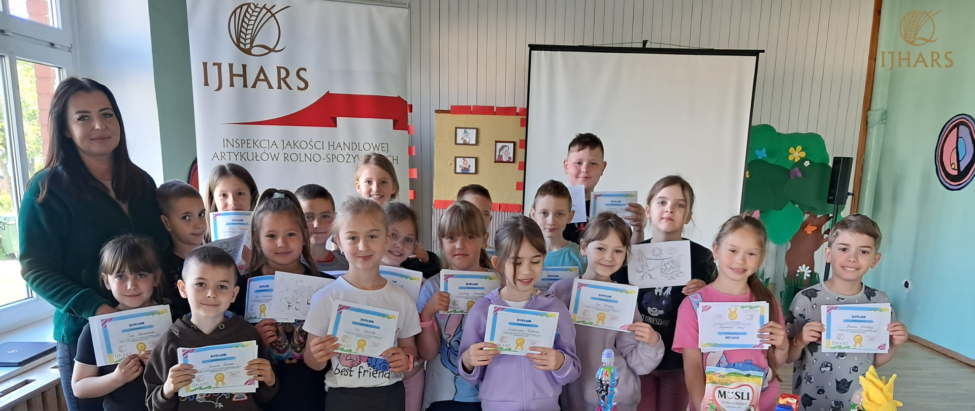 Zdjęcie grupowe dzieci po zakończonej lekcji wraz z Inspektorką IJHARS. Uczestnicy stoją uśmiechnięci w sali edukacyjnej, a w tle widoczne jest logo IJHARS. Atmosfera przyjazna i edukacyjna.