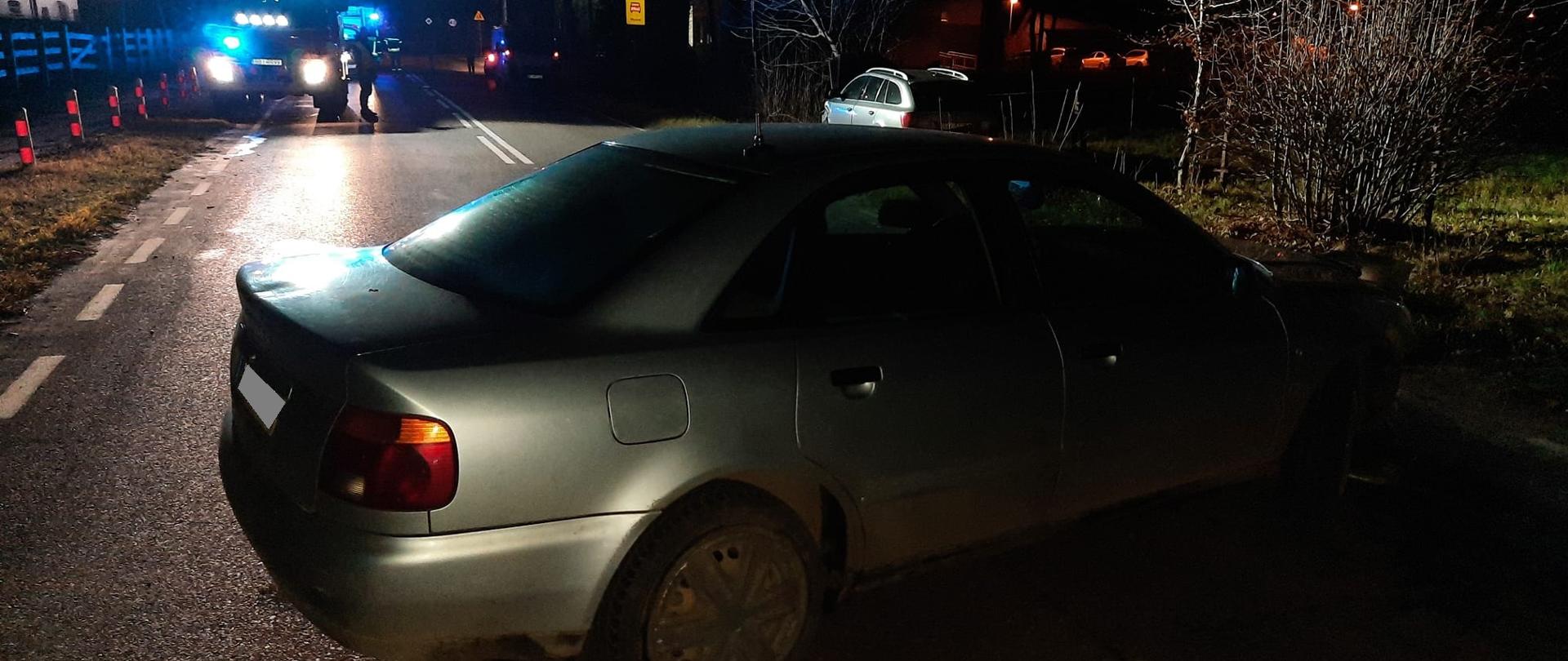 Samochód osobowy po zderzeniu z innym pojazdem stojący w poprzek jezdni. W tle drugi z pojazdów biorących udział w zdarzeniu stojący na poboczu oraz samochód strażacki.