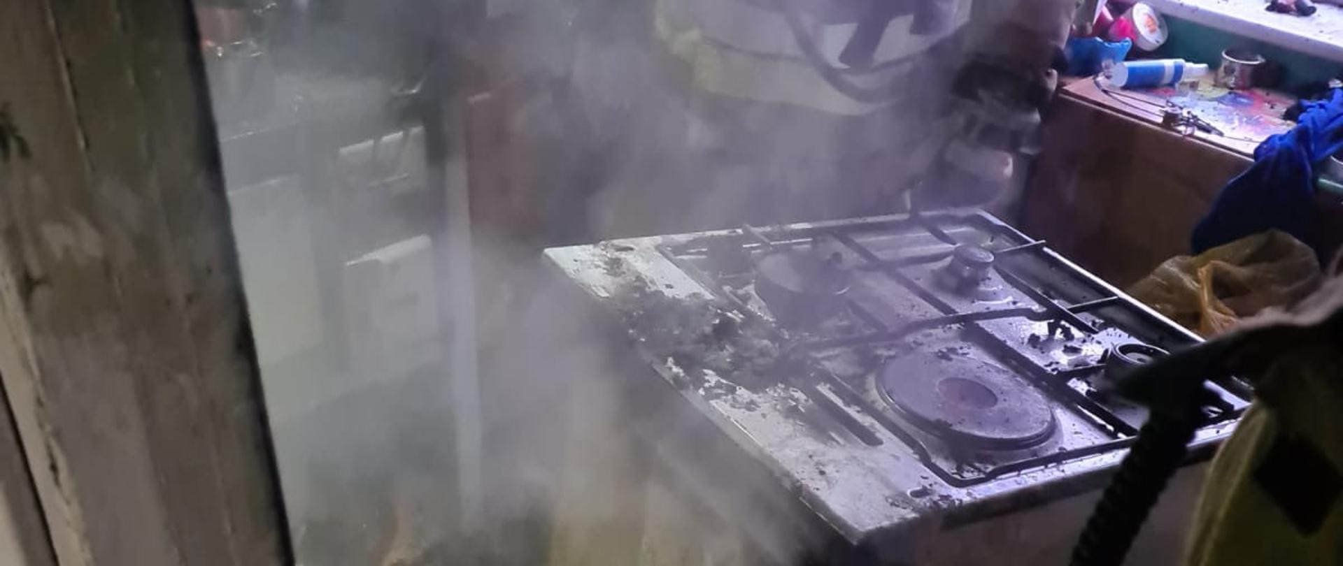 Fotografia przedstawia zbliżenie na wnętrze pomieszczenia w którym wybuchł pożar. W centralnej części kadru widoczna kuchenka gazowa, obok kuchenki po jej obu stronach widoczni strażacy w ubraniach specjalnych. W pomieszczeniu unosi się jeszcze śladowa ilość dymu, na podłodze widoczne zwęglone pozostałości po pożarze.