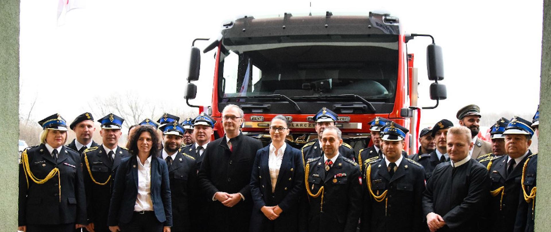 Przekazanie nowych samochodów pożarniczych - strażacy ubrani w mundury wyjściowe stoją na tle nowego samochodu ratowniczo-gaśniczego
