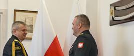 Pomorski komendant wojewódzki Państwowej Straży Pożarnej podaje dłoń strażakowi, mężczyźni stoją na tle flag państwowych Polski.