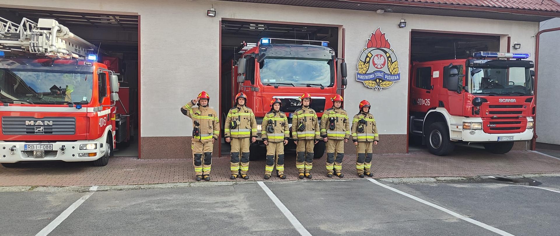 Na zdjęciu widać garaże Jednostki Ratowniczo Gaśniczej w Ustrzykach Dolnych, plac zewnętrzny, pojazdy ratowniczo gaśnicze z włączonymi sygnałami świetlnymi oraz sześciu strażaków PSP w ubraniach specjalnych. Dowódca stojący jako pierwszy od lewej oddaje honor. 