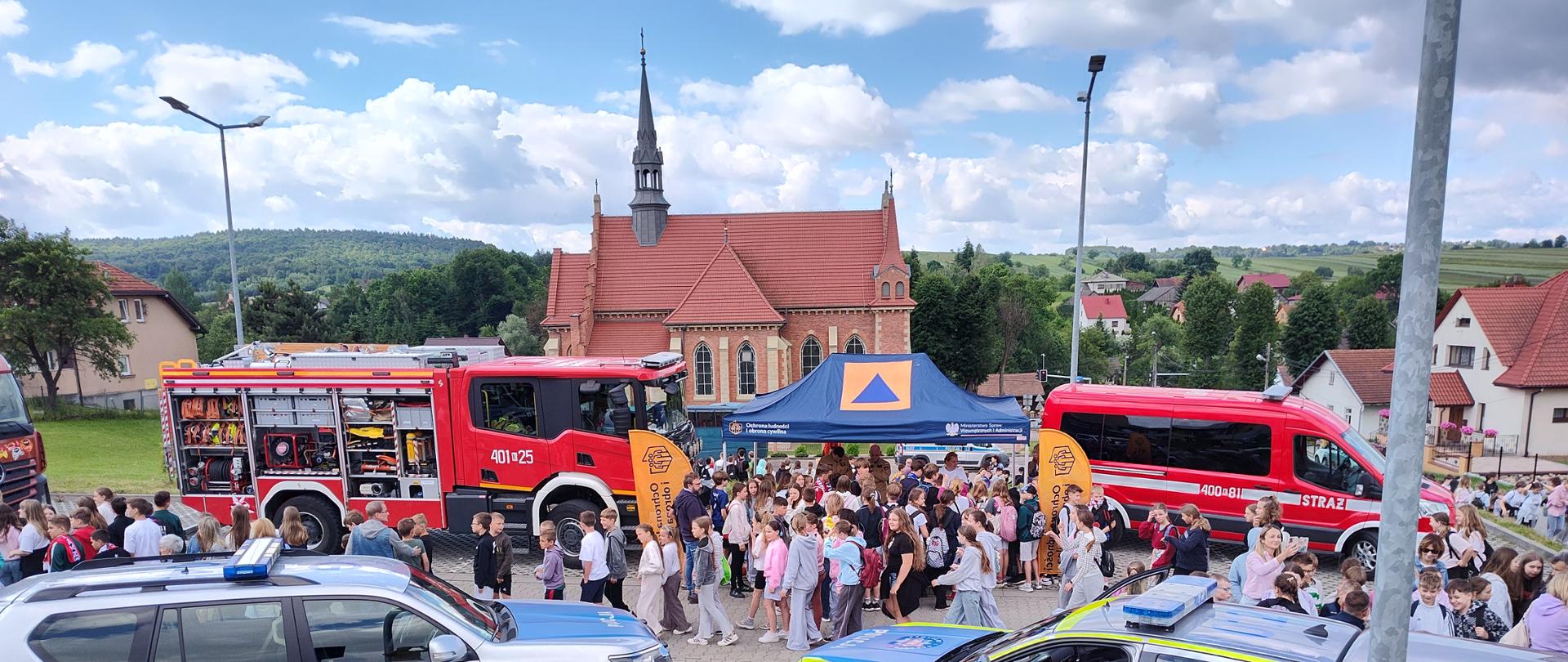 Zdjęcie wykonane z podwyższenia, przedstawiające plac manewrowy z ułożonych betonowych płyt ażurowych, na którym zorganizowano pokazy służb ratunkowych. Na pierwszym planie stoją dwa srebrno-niebieskie radiowozy policyjne. W tle widać czerwony wóz strażacki z otwartymi skrytkami, granatowy namiot edukacyjny otoczony przez tłum dzieci oraz czerwony mikrobus strażacki. W tle widoczny jest zabytkowy kościół z cegły oraz zielone wzgórza.