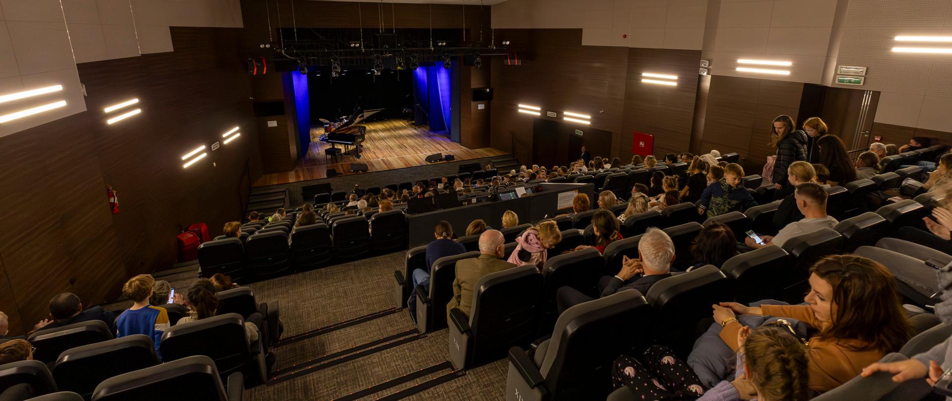 scena w domu kultury w głubczycach, zdjęcie wykonane z punktu bardzo oddalonego od sceny, perspektywa widza siedzącego na tyle sali, widoczne osoby siedzące na publicznoś