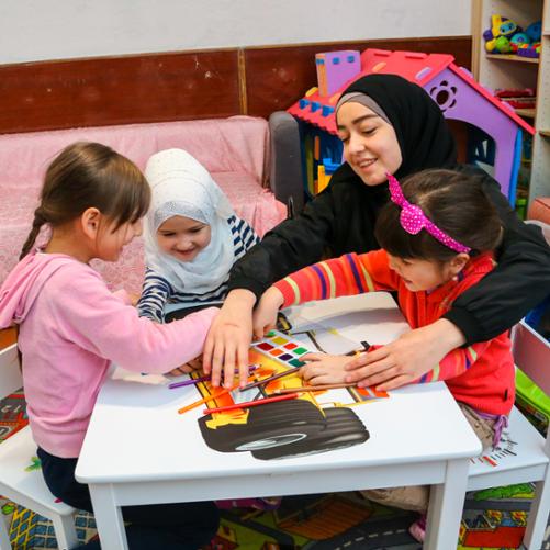 uśmiechnięta kobieta siedzi przy stole z trzema dziewczynkami i rysują na stoliku 