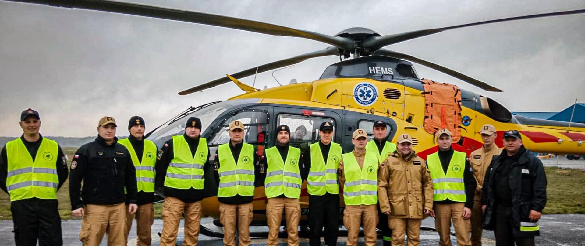 Strażacy pozują do zdjęcia grupowego na tle żółtego śmigłowca LPR, który stoi na płycie lotniska. 