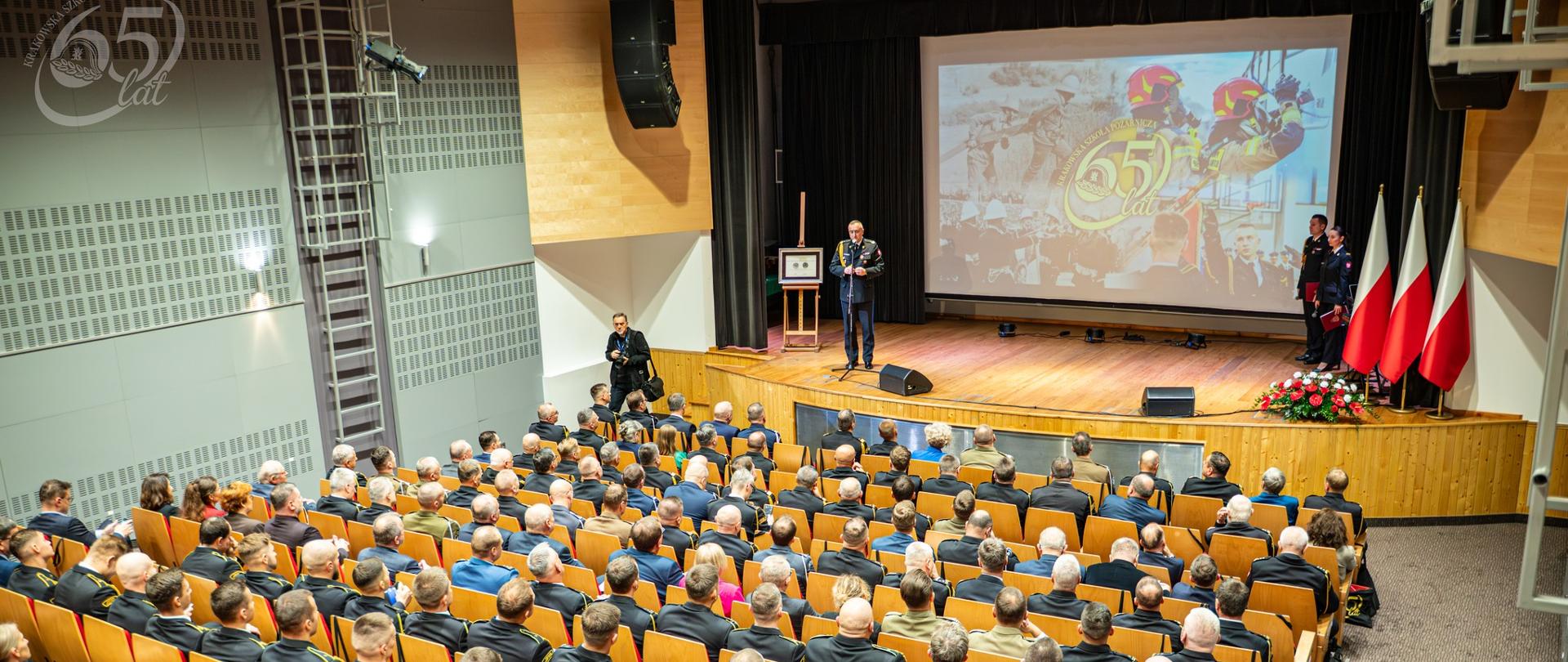 4 Widok Ogólny na Salę Audytoryjną
Szerokie ujęcie sali audytoryjnej. Na drewnianej scenie, w centrum, stoi pojedynczy mówca przy mównicy. Po prawej stronie sceny stoją cztery flagi Polski z czerwoną i białą tkaniną ułożone obok siebie, u podstawy mają kompozycję z czerwonych róż. Publiczność, składająca się z kilkuset osób, siedzi tyłem w rzędach krzeseł. Większość uczestników jest w ciemnych mundurach galowych lub służbowych. W tle sceny widoczny jest duży ekran projekcyjny z barwną grafiką "65 lat".
