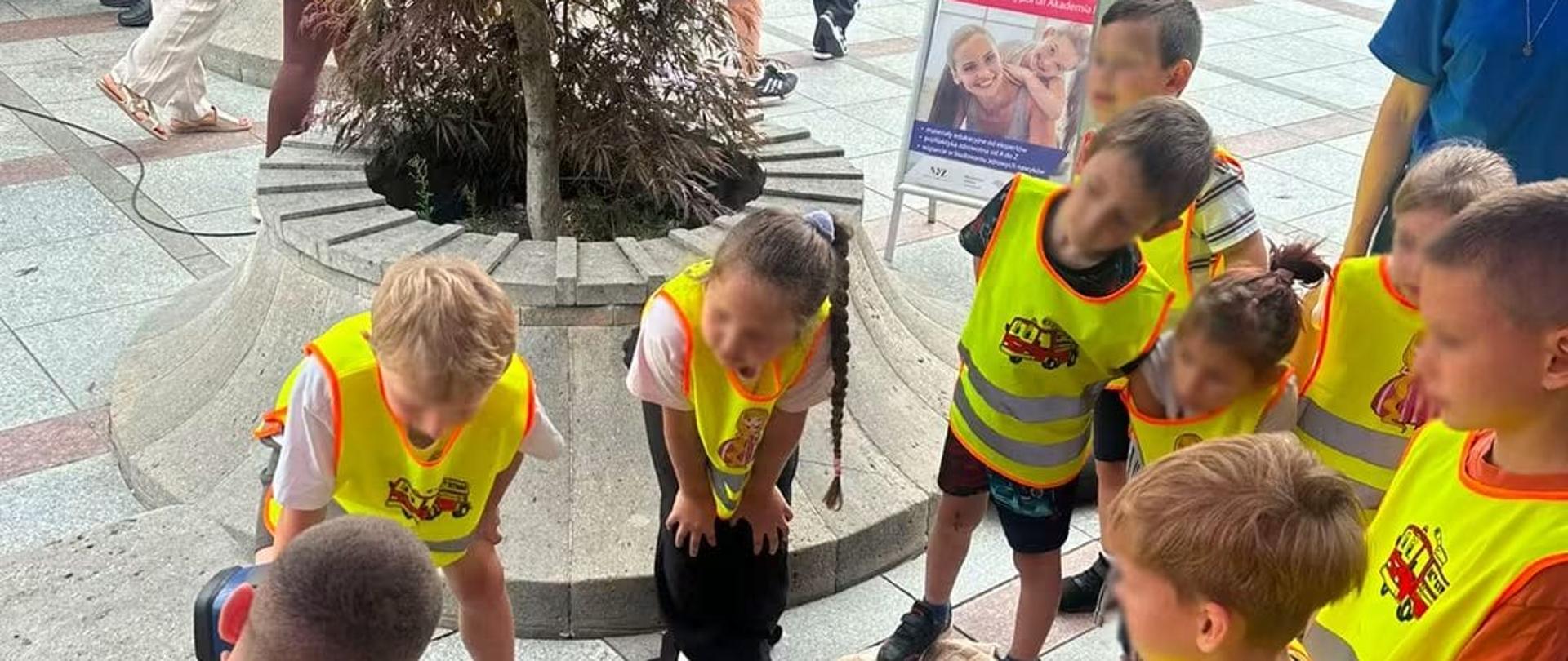 Strażak pod namiotem edukacyjnym prezentuje dzieciom w kamizelkach odblaskowych i ich opiekunom strój strażacki oraz sprzęt ratowniczy ułożony na stole. Przed stoiskiem leży manekin do resuscytacji oraz torby ze sprzętem medycznym.