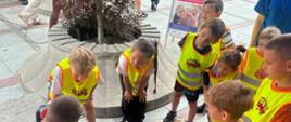 Strażak pod namiotem edukacyjnym prezentuje dzieciom w kamizelkach odblaskowych i ich opiekunom strój strażacki oraz sprzęt ratowniczy ułożony na stole. Przed stoiskiem leży manekin do resuscytacji oraz torby ze sprzętem medycznym.