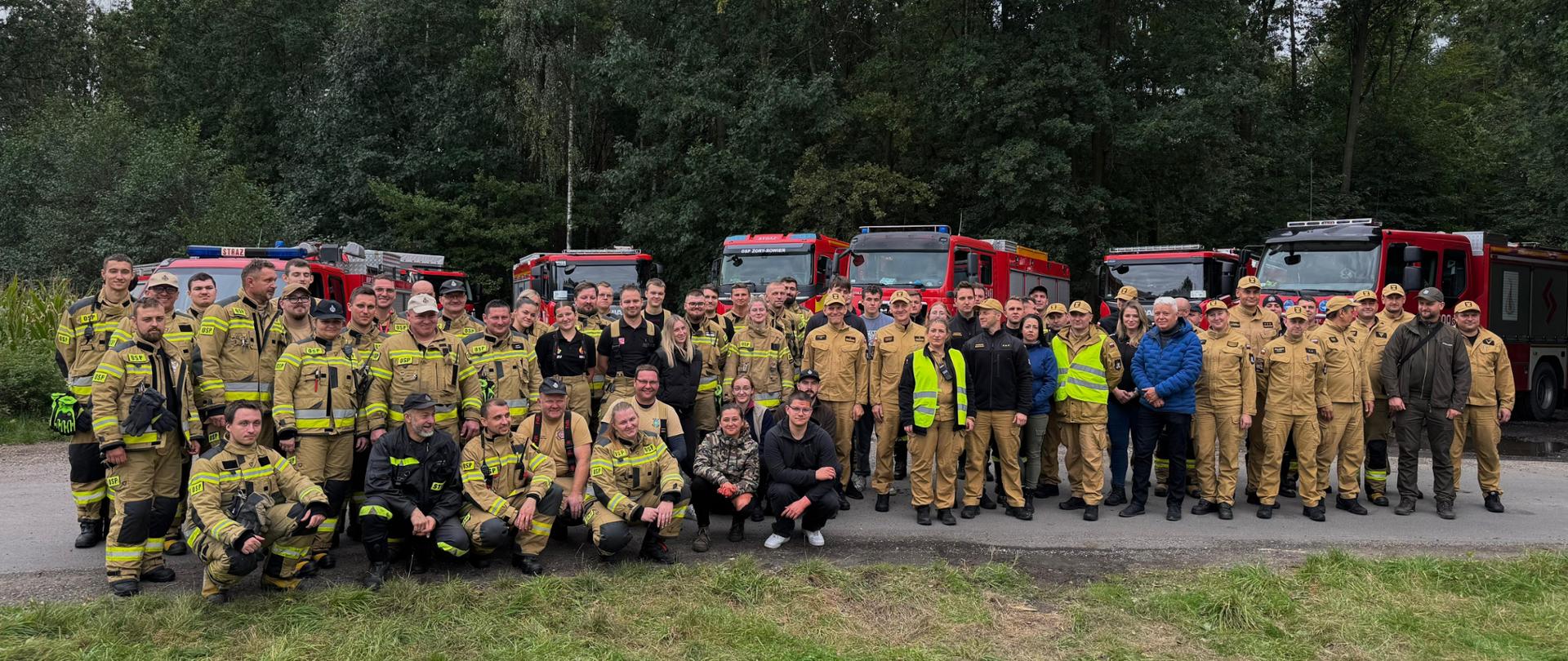 Grupa strażaków i ratowników w różnych mundurach, ustawiona przed szeregiem czerwonych wozów strażackich na tle zieleni i lasu.
