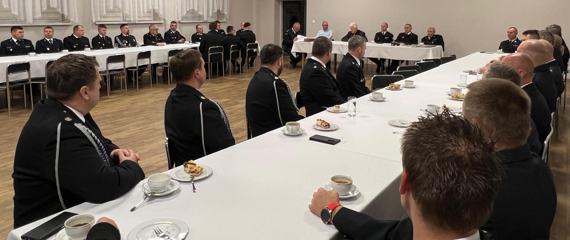 Na zdjęciu widać formalne spotkanie z udziałem dużej grupy osób. Uczestnicy: Większość osób ma na sobie ciemne, galowe mundury, co silnie sugeruje, że są to funkcjonariusze straży pożarnej, na co wskazują dystynkcje na pagonach i odznaczenia widoczne na kurtkach). Wśród siedzących jest widoczna co najmniej jedna kobieta w mundurze. Stoły: Uczestnicy siedzą przy dwóch długich stołach przykrytych białymi obrusami. Stoły są ustawione w kształcie litery "U".
Nakrycie: Na stołach znajdują się liczne białe filiżanki z ciemną obwódką i spodeczkami, najprawdopodobniej z kawą lub herbatą. Widać też małe talerzyki oraz sztućce.
Lokalizacja: Spotkanie odbywa się w przestronnej, jasnej sali o charakterze konferencyjnym lub bankietowym.
Tło: Na ścianie widoczne są okna zasłonięte jasnymi, marszczonymi firanami z koronkowym wykończeniem.
Sufit: Sufit jest podwieszany, z wbudowanymi prostokątnymi oprawami oświetleniowymi
Podłoga: Na podłodze widoczne są panele.