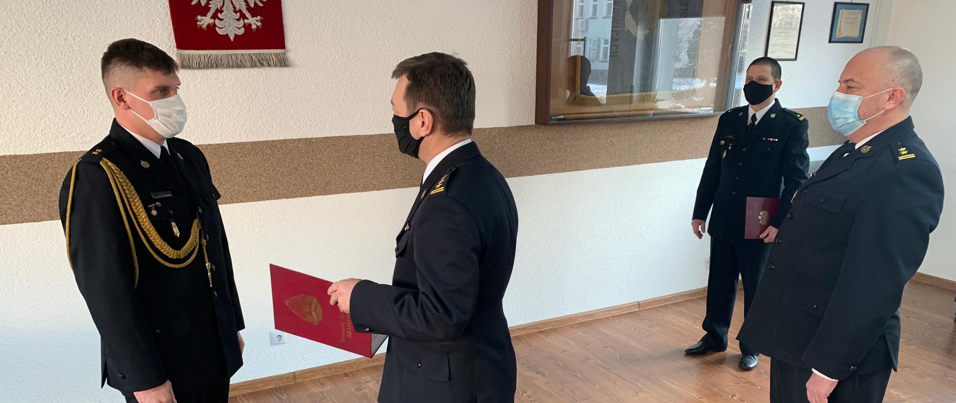 Zdjęcie zrobione na świetlicy Komendy powiatowej PSP w Krapkowicach przedstawia moment wręczenia decyzji o mianowaniu na stanowisko zastępcy dowódcy JRG. Oficerowie w mundurach wyjściowych. Od lewej stoi oficer mianowany – mł. kpt. Przemysław Zając, a naprzeciw niego Komendant Powiatowy st. bryg. Krzysztof Kuniec z teczką koloru czerwonego zawierającą decyzję. Po prawej za komendantem stoi jego zastępca – bryg. Rafał Bisowski, a w tle dowódca JRG – mł. bryg. Lucjan Lubaszka. Tło stanowi biała ściana z godłem narodowym oraz gablotą ze sztandarem. W związku z ograniczeniami związanymi zapobieganiu rozprzestrzeniania COVID-19 strażacy na twarzach mają założone maseczki ochronne. 