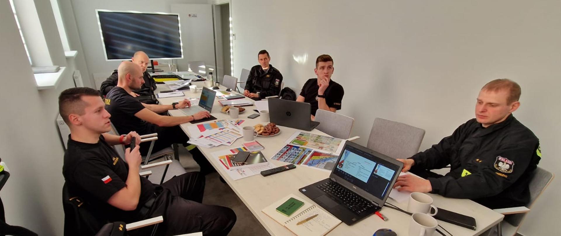 strażacy siedzący przy stole, na środku duży telewizor ze slajdem, na stole dużo dokumentów i komputer