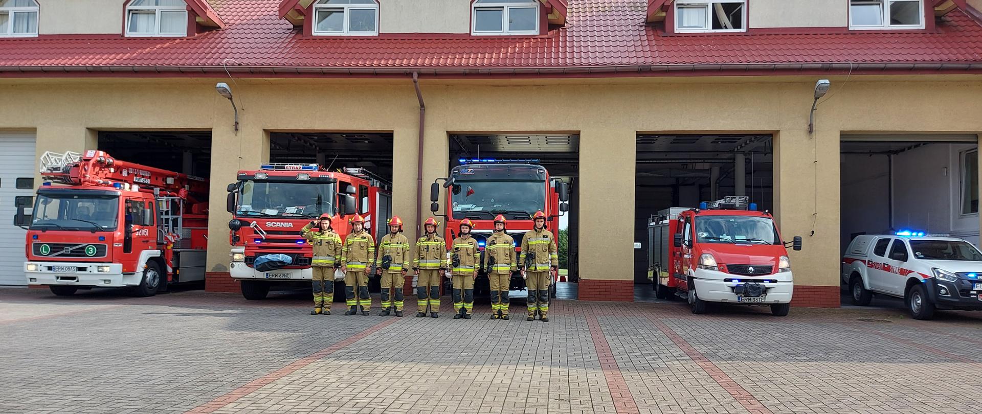 Zdjęcie przedstawia 5 otwartych drzwi garażowych straży pożarnej, w których soją samochody strażackie, a przed nimi w jednym szeregu ustawionych jest 7 strażaków, ubranych w piaskowe stroje bojowe, z hełmami na głowie.