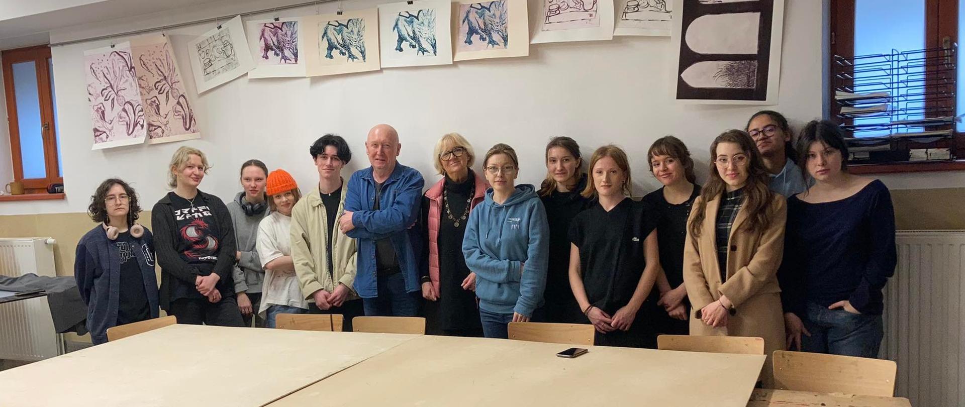 Na zdjęciu występują uczestnicy warsztatów Mokulito. Na pierwszym planie widzimy duży stół. Za stołem pod ścianą znajduje się grupa uczniów wraz z prowadzącymi warsztaty: profesorem Józefem Budką i jego małżonką profesor Aleksandrą Budką. W grupie młodzieży jest również pani Agnieszka Rokicka - wicedyrektor PLSP im. Antoniego Kenara.