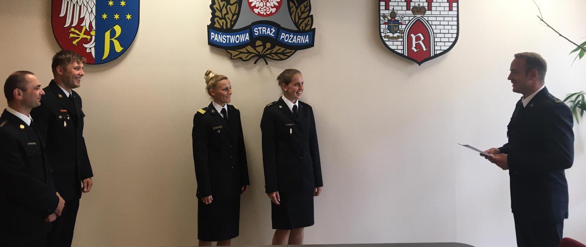 Zdjęcie wykonane w pomieszczeniu Komendy Miejskiej Państwowej Straży Pożarnej w Radomiu. Na zdjęciu widać po prawej stronie Zastępcę Komendanta Miejskiego Państwowej Straży Pożarnej trzymającego w rękach akty mianowania na wyższe stanowiska służbowe. Po środku stoją dwie funkcjonariuszki, które otrzymają akty. Po lewej stronie na zdjęciu stoją dwaj funkcjonariusze – bezpośredni przełożeni osób mianowanych. Wszyscy funkcjonariusze są ubrani w umundurowanie galowe. Nad nimi na ścianie znajdują się herb powiatu radomskiego, logo Państwowej Straży Pożarnej oraz herb miasta Radomia. 