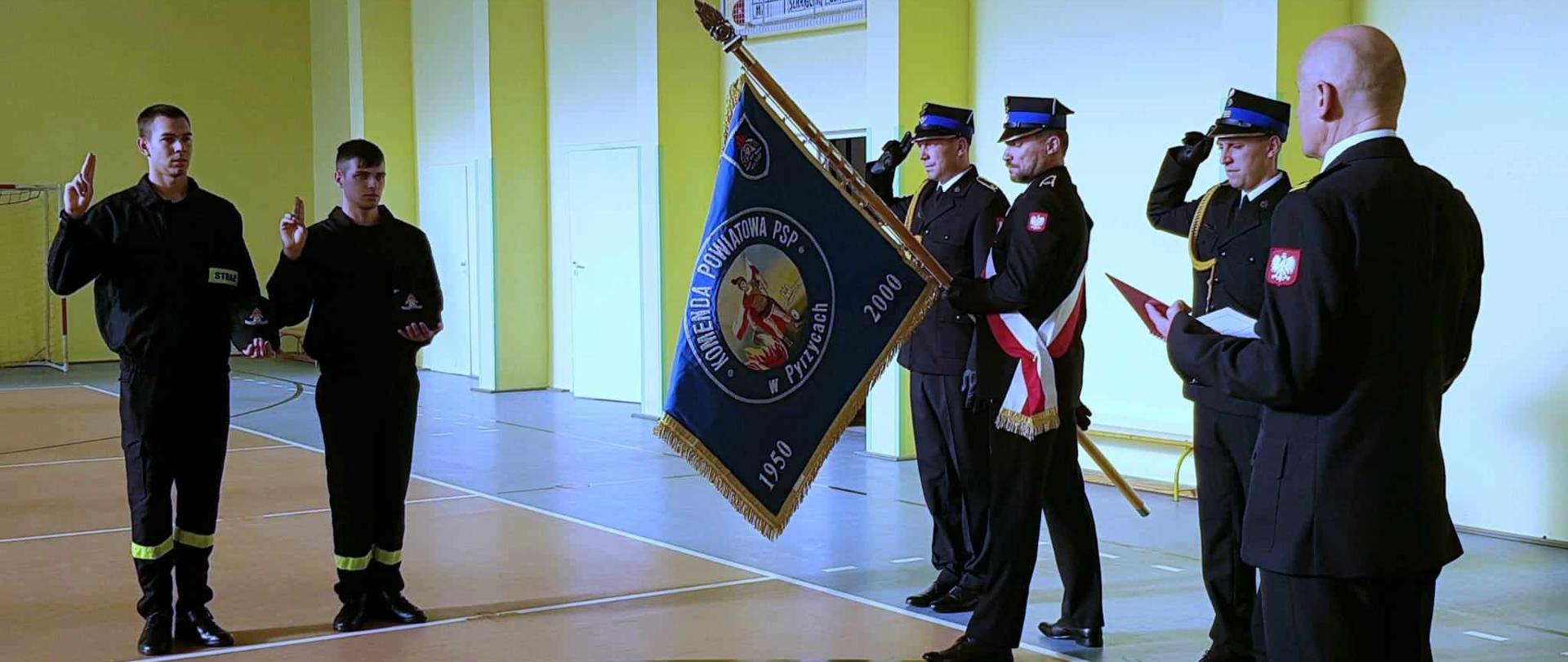 Dwóch strażaków składa rotę ślubowania w obecności komendanta i pocztu sztandarowego