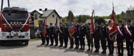 Z lewej strony zdjęcia stoi samochód ratowniczo-gaśniczy, po prawej stronie stoją ustawione w rzędzie poczty sztandarowe.