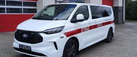 Na zdjęciu widać biały samochód straży pożarnej przez który przebiega czerwonym pasem z napisem „STRAŻ”. Samochód jest zaparkowany przed budynkiem z czerwonymi bramami garażowymi. Budynek ma kilka okien dachowych.