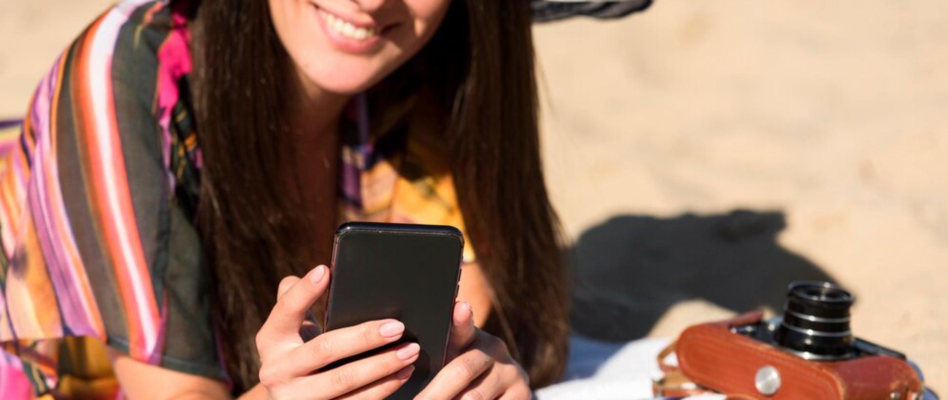 Kobieta w kapeluszu na plaży trzymająca smartfon. Obok na ręczniku leży aparat fotograficzny i okulary przeciwsłoneczne 