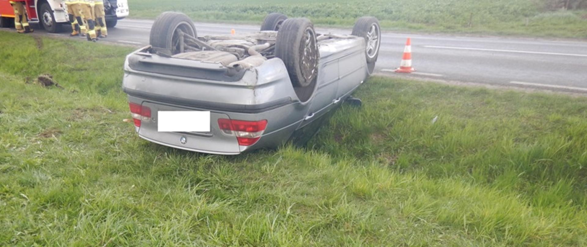 Zdjęcie przedstawia srebrny samochód osobowy po wypadku drogowym. Samochód leży na dachu.