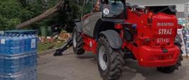 Na zdjęciu widoczna jest ładowarka teleskopowa Manitou wykorzystywana przez strażaków podczas usuwania powalonego drzewa. W tle widoczny jest uszkodzony konar oraz teren prowadzonych działań ratowniczych.