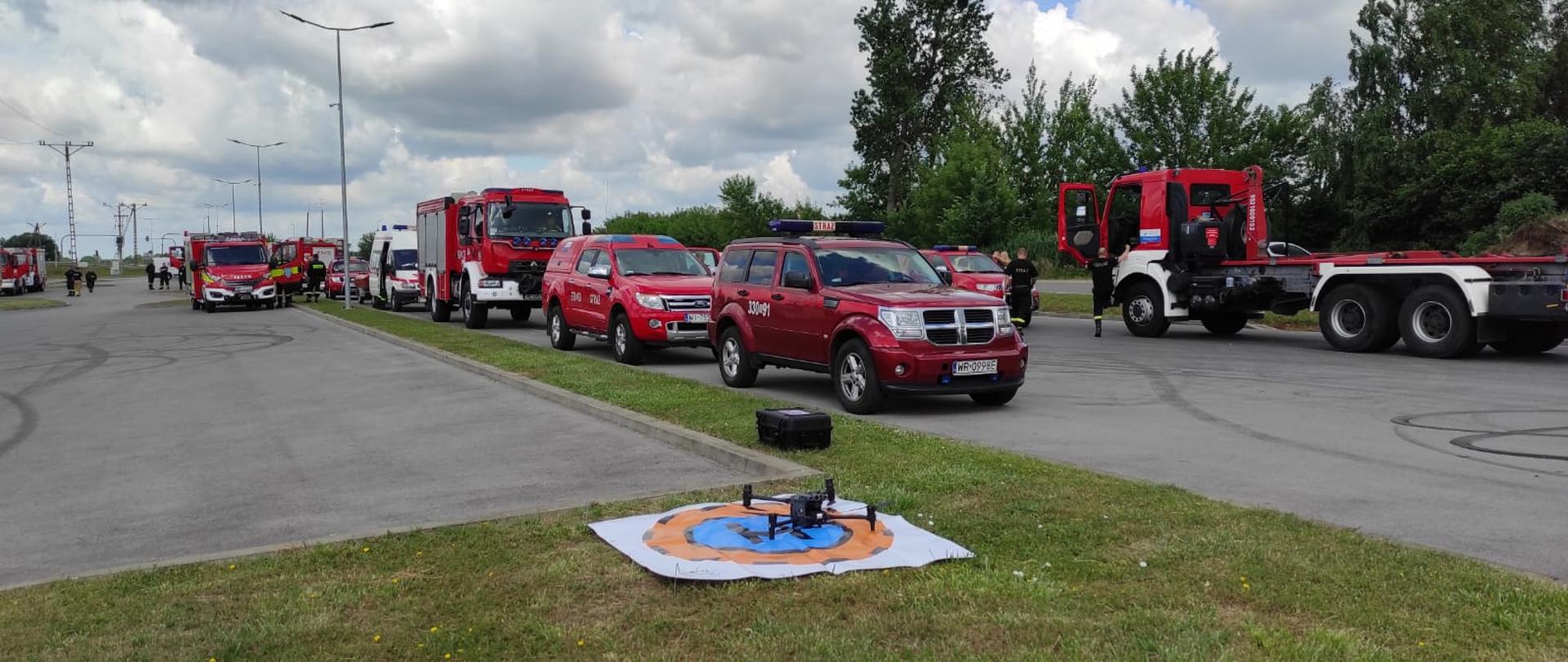Zdjęcie wykonane podczas ćwiczeń w miejscowości Jedlińsk w pobliżu rzeki Radomka. Na zdjęciu widać pojazdy pożarnicze biorące udział w ćwiczeniach. Pojazdy terenowe oraz ratowniczo – gaśnicze. Pojazdy w kolorze czerwonym z białymi oraz srebrnymi elementami zabudowy. Przed nimi na trawie na specjalnej macie ustawiony jest dron. 