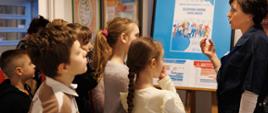 Grupa dzieci stoi i słucha prezentacji prowadzonej przy planszy informacyjnej. Na planszy widoczny jest plakat dotyczący szczepień ochronnych, a osoba prowadząca wskazuje na jego treść. W tle znajdują się tablice i gazetki edukacyjne.
