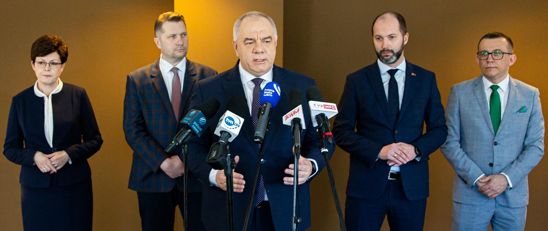 Na zdjęciu wicepremier Jacek Sasin stoi za mikrofonami. Za nim stoją pozostali uczestnicy - trzech mężczyzn i kobieta. 