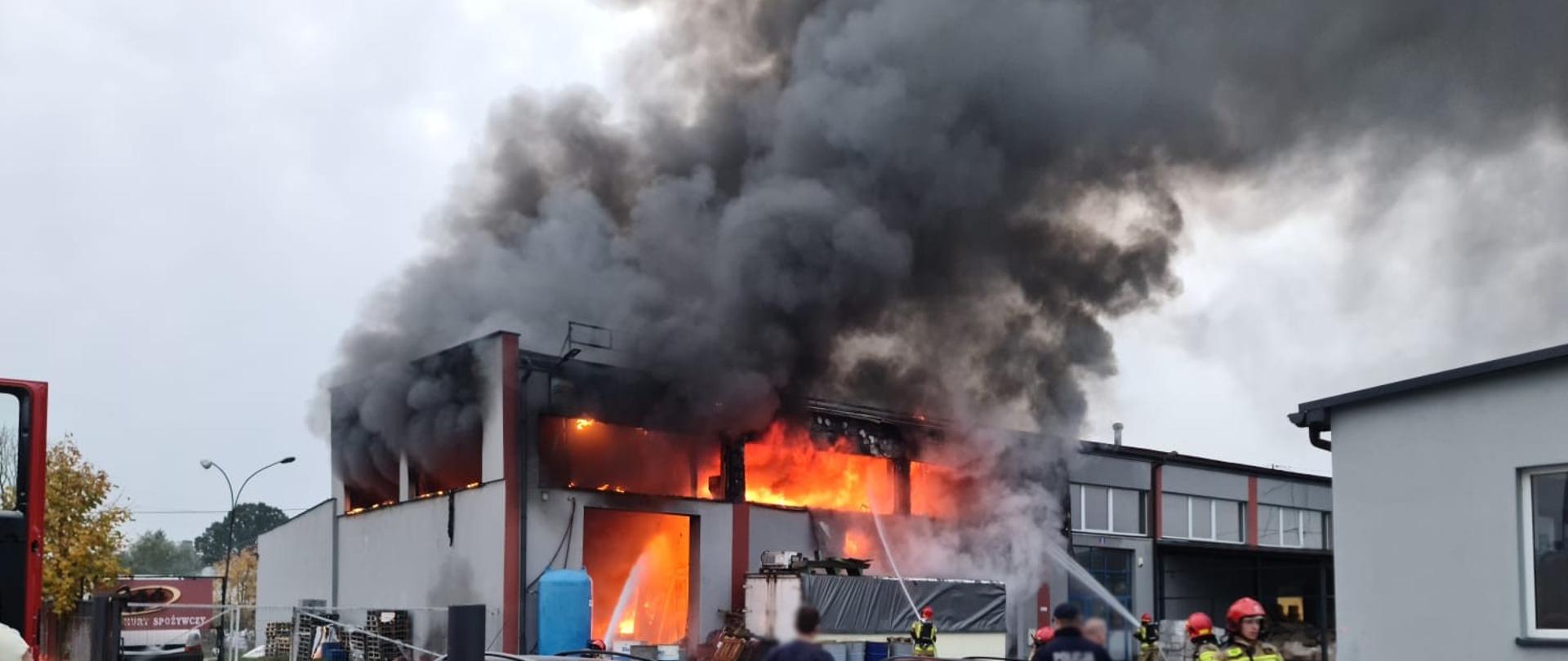 Płomienie i dymy wydobywające się z hali a obok ludzie oraz strażacy.